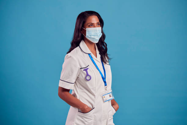 Portrait Of Female Mature Nurse In Uniform Wearing Face Mask In Front Of Blue Studio Background stock photo