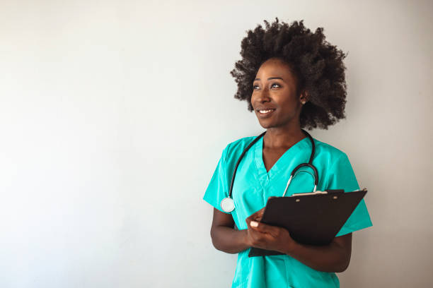 Portrait Of Female Nurse Standing In Hospital Corridor Female nurse or doctor smiles while staring out window in hospital hallway and holding clipboard with patient file. African American female pediatric nurse in office. Portrait Of Female Nurse Standing In Hospital Corridor female nurse stock pictures, royalty-free photos & images