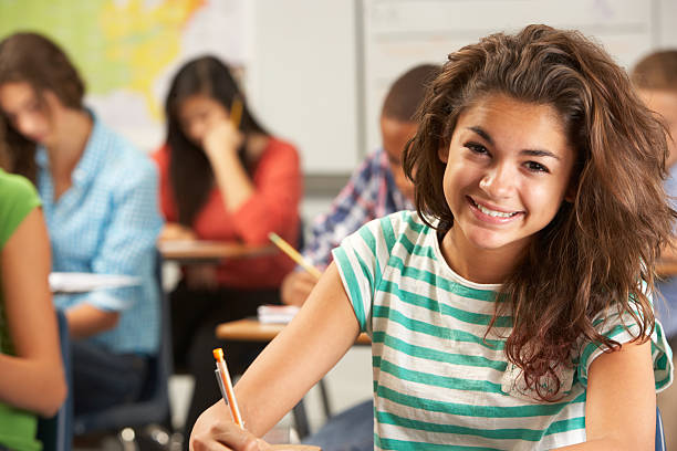 Portrait Of Female Pupil Studying At Desk In Classroom Portrait Of Female Pupil Studying At Desk In Classroom Smiling At Camera latina girl stock pictures, royalty-free photos & images