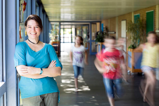 portrait of female teacher, leaning at corridor wall, running children in background stock photo