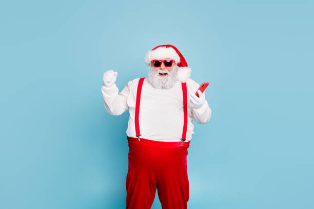 Portrait of funny funky crazy christmas father with big belly use mobile phone raise fists scream yes win in x-mas winter shopping lottery wear red suspenders isolated over blue color background Portrait of funny funky crazy christmas father with big belly use mobile phone raise, fists scream yes win in x-mas winter shopping lottery wear red suspenders isolated over blue color background fat man looks at the phone stock pictures, royalty-free photos & images