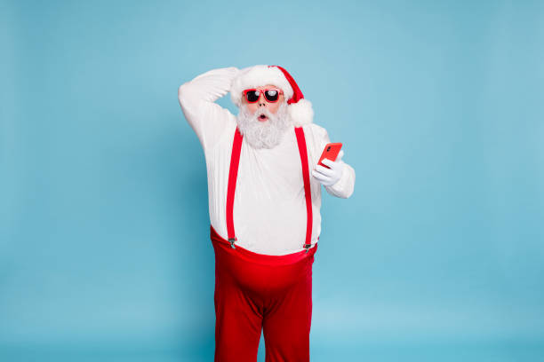 Portrait of funny funky fat modern santa claus in sunglass use his cell phone got christmas time blogs notification scream wow omg wear red suspenders overalls isolated over blue color background Portrait of funny funky fat modern santa claus in sunglass use his cell phone got christmas time blogs notification scream wow omg wear red suspenders, overalls isolated over blue color background fat man looks at the phone stock pictures, royalty-free photos & images