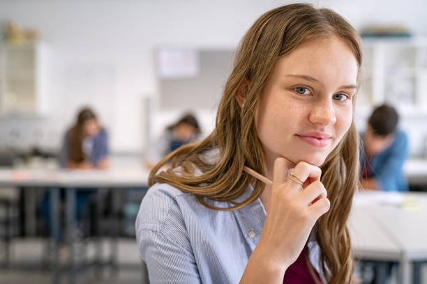 Portrait of girl in high school classroom Portrait of college student sitting at desk holding pencil near chin while looking at camera. Portrait of high school girl studying with classmates in library with copy space. Close up face of young smart woman in casual with hand on chin thinking. 16 17 years stock pictures, royalty-free photos & images