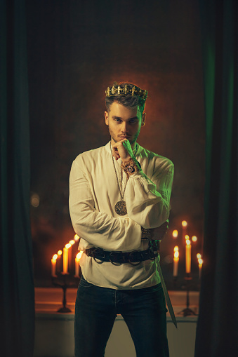 Portrait of handsome attractive man in image of medieval king. Vintage clothing, historic white shirt retro clothing gold crown on head. Dark gothic room, studio. Fashion model. Adult sexy guy knight