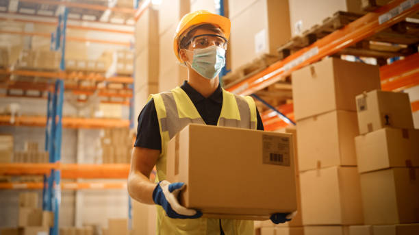 Portrait of Handsome Male Worker Wearing Medical Face Mask and Hard Hat Carries Cardboard Box Walks Through Retail Warehouse full of Shelves with Goods. Safety First Protective Workplace.  storage boxes stock pictures, royalty-free photos & images