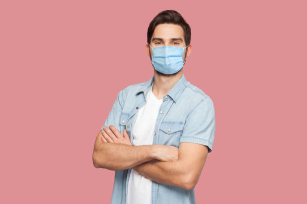 porträtt av stilig ung man med kirurgisk medicinsk mask i blå casual stil skjorta stående, upphöjda armar och titta på kameran med leende. - face mask bildbanksfoton och bilder