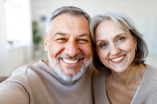 Portrait of happy beautiful senior caucasian family couple smiling at camera, cheerful retired wife and husband making selfie on smartphone while spending time together at home