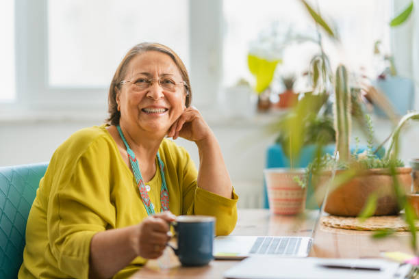 Portrait of happy fashionable senior woman smiling for camera while using laptop at home stock photo