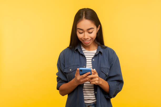 Portrait of happy girl in stylish denim shirt typing message and smiling, dialing number calling on cell phone Portrait of happy girl in stylish denim shirt typing message and smiling, dialing number calling on cell phone, using online mobile application, searching web. indoor studio shot, yellow background indonesian girl stock pictures, royalty-free photos & images