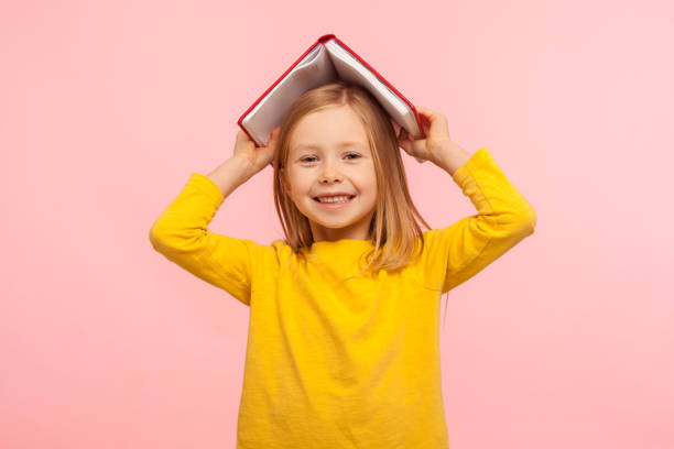 portret van gelukkig lui meisje dat hoofd met boek behandelt en aan camera glimlacht, ongehoorzaam kind dat pret heeft - portrait girl stockfoto's en -beelden