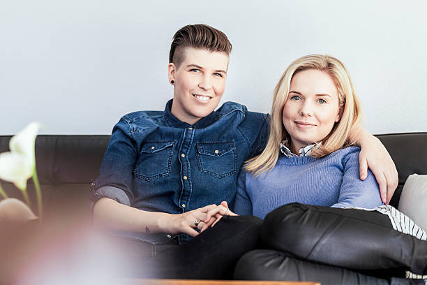 Portrait of happy lesbian couple sitting on sofa Portrait of happy lesbian couple sitting on sofa. Relaxed young homosexual partners are in living room. Smiling women are at home. gay couple ring tattoos stock pictures, royalty-free photos & images