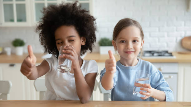Portrait of happy multiracial children enjoying drinking water. Portrait of happy pretty little thirsty african ethnicity kid girl drinking pure stilled water, showing thumbs up gesture with smiling small european friend or foster sister, sitting in kitchen. drinking water stock pictures, royalty-free photos & images