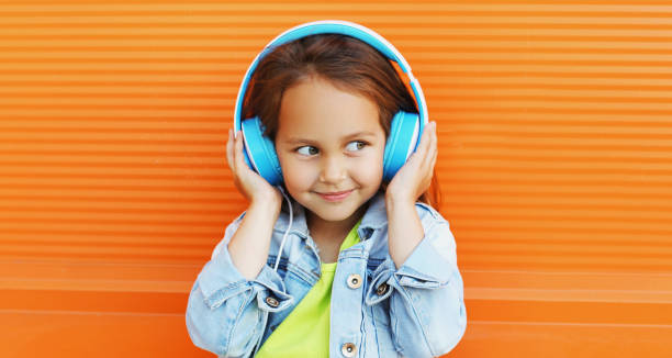 Portrait of happy smiling child in headphones listening to music on city street over orange wall background stock photo