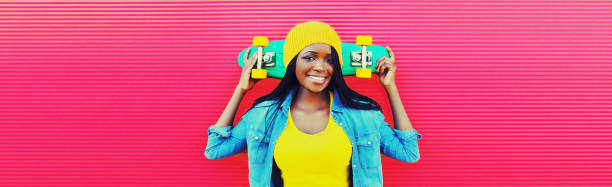 Portrait of happy smiling young african woman with skateboard wearing colorful clothes, hat on vivid background stock photo