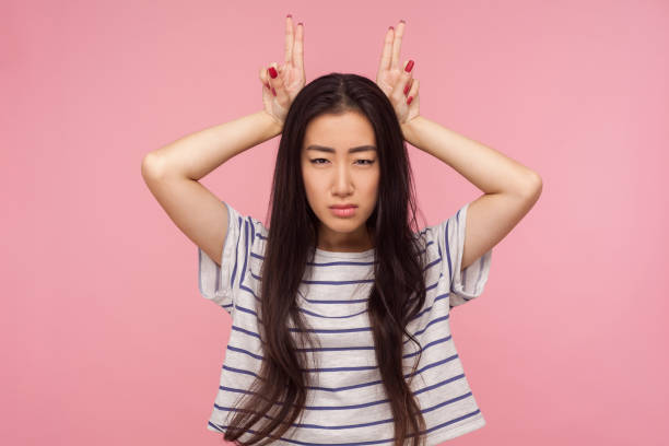 Portrait of irritated girl in striped t-shirt threatening with bull horn gesture, looking at camera with hostile expression Portrait of irritated girl in striped t-shirt threatening with bull horn gesture, looking at camera with hostile suspicious expression, ready to attack. indoor studio shot isolated on pink background ugly girl stock pictures, royalty-free photos & images