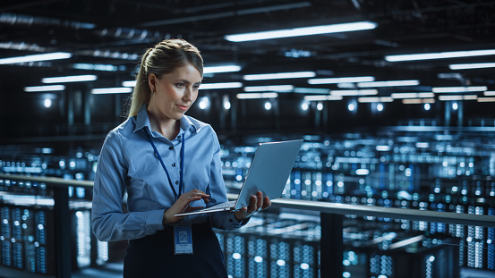 Portrait of IT Specialist Using Laptop in Data Center. Big Server Farm Cloud Computing Facility with Female System Administrator Monitoring. Cyber Security, Network Protection Services.