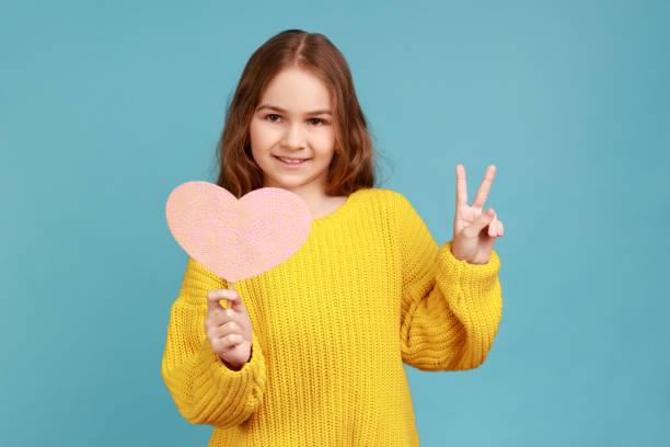 Potret gadis kecil memegang kertas hati merah muda di tongkat dan tersenyum ke kamera, menunjukkan tanda v. foto stok