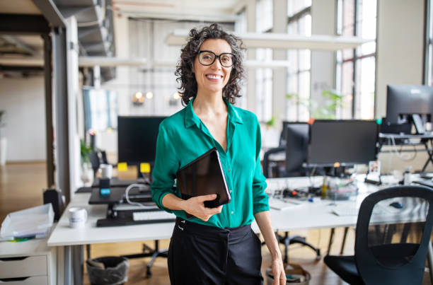 Portrait of mature businesswoman in casuals Portrait of mature businesswoman in casuals standing at her desk with digital tablet. Woman entrepreneur in office looking at camera and smiling. business casual photos stock pictures, royalty-free photos & images