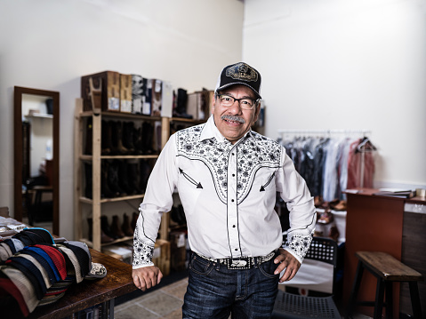 Portrait of mature Latin man dressed as Vaquero-Mexican Cowboy. He has grey moustache, wearing eyeglasses and western themed clothes with cowboy hat, shirt and jeans. Interior of Mexican Cowboy retail store.