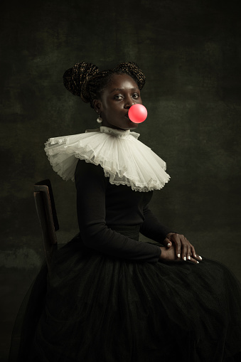 Bubbles gum. Medieval African young woman in black vintage dress with big white collar isolated on dark green background. Concept of comparison of eras, modernity and renaissance. White pearls
