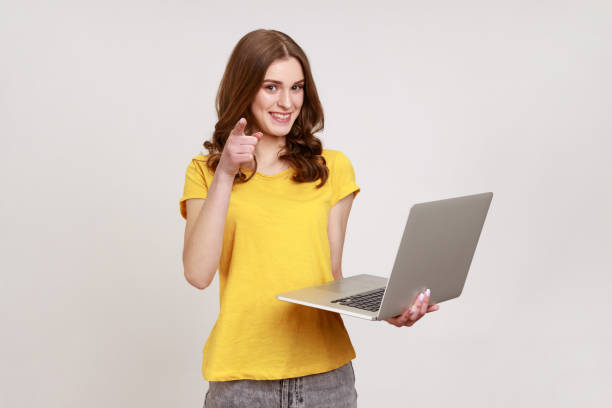 Portrait of optimistic young woman in yellow T-shirt holding laptop, pointing finger to camera and smiling, choosing you, freelance job advertisement. stock photo