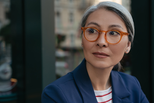Close up portrait of pensive mature businesswoman looking away, planning start up. Beautiful asian woman wearing stylish eyeglasses standing outdoors, focus on face