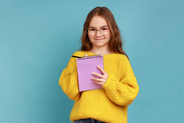 Portrait of positive little girl writing in notebook, looks smiling at camera, child doing homework. stock photo