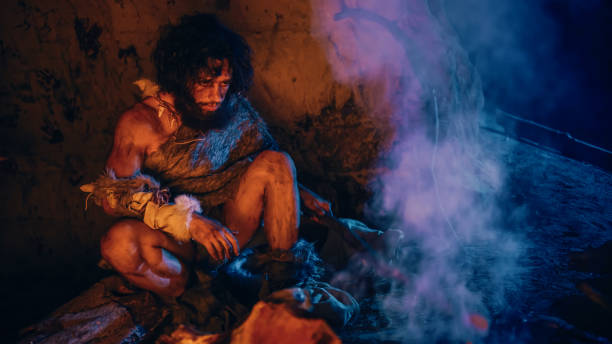 portrait of primeval caveman wearing animal skin sitting in his cave, warms up near the fire. primitive neanderthal hunter / homo sapiens at night alone in his den - fire caveman imagens e fotografias de stock