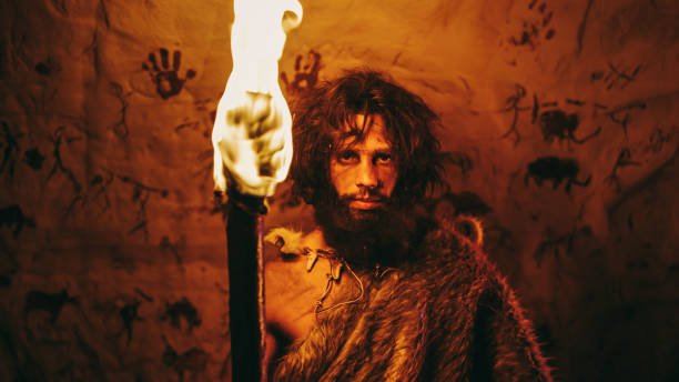 portrait of primeval caveman wearing animal skin standing in his cave at night, holding torch with fire. primitive neanderthal hunter / homo sapiens at night alone. in the background cave art drawings - fire caveman imagens e fotografias de stock
