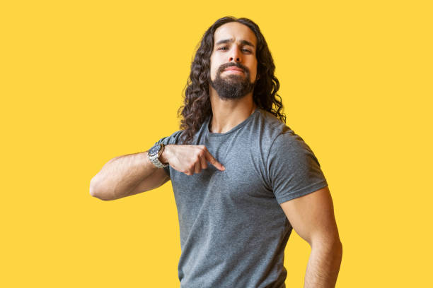 Portrait of proud bearded young man with long curly hair in grey tshirt standing, pointing himself and looking at camera with confident serious face. Portrait of proud bearded young man with long curly hair in grey tshirt standing, pointing himself and looking at camera with confident serious face. indoor studio shot isolated on yellow background. arrogance stock pictures, royalty-free photos & images