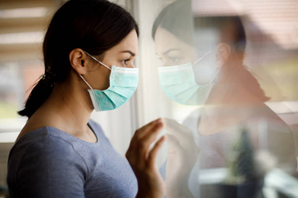 Portrait of sad young woman with face protective mask looking through the window at home Portrait of sad young woman with face protective mask looking through the window at home quarantine stock pictures, royalty-free photos & images