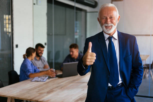 Portrait of senior businessman showing thumb up and smiling with group of people in the background Portrait of senior businessman showing thimbs up with group of business people in the background business thumbs up stock pictures, royalty-free photos & images