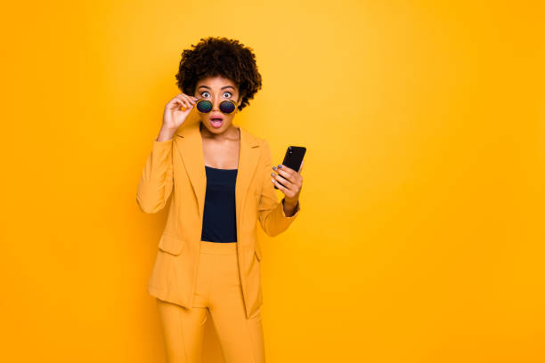 porträtt av chockade afro american girl använda smartphone läs nyheter på sociala nätverkskonto få meddelande om ogillar scream omg slitage snygga moderiktiga kläder isolerade gul färg bakgrund - omg girl bildbanksfoton och bilder