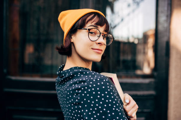 portret van slimme jonge vrouwelijke student dragen gele hoed, transparante brillen en groen met witte stippen shirt met boeken in handen staan tegen de universiteit. - student stockfoto's en -beelden