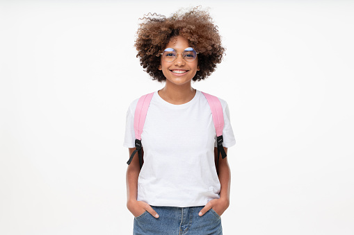 Portrait of smiling african school girl wearing white t-shirt, glasses and backpack, isolated on gray background