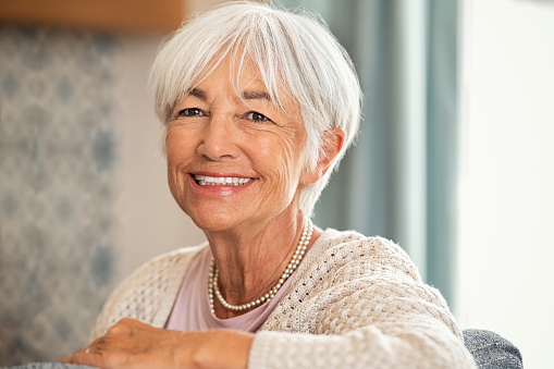 Happy old woman with white hair sitting on sofa and looking at camera. Retired woman relaxing on her couch in the living room. Close up face of cheerful senior lady with big grin enjoying retirement.