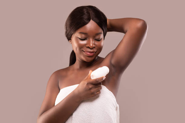 Portrait of smiling black woman applying roller deodorant to armpit zone Portrait of smiling black woman applying roller deodorant to armpit zone. Beautiful african american lady using antiperspirant stick for underarms standing wrapped in towel over gray background deodorant stock pictures, royalty-free photos & images