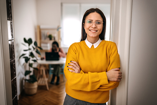 Portrait of smiling young businesswoman wearing yellow sweatshirt standing at entrance in small office