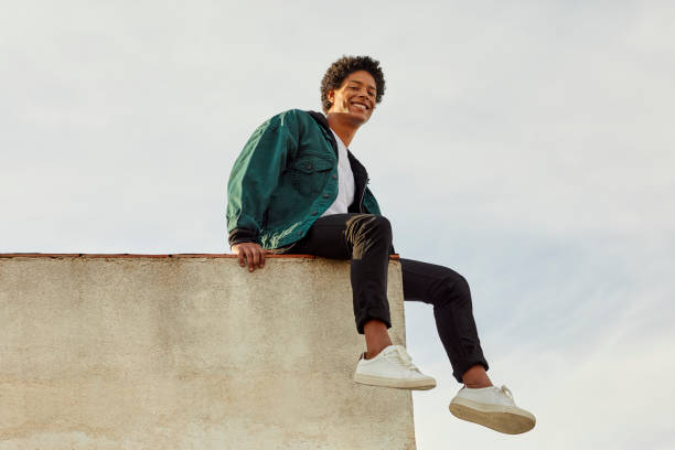 Portrait of smiling carefree man at rooftop Portrait of smiling man sitting on retaining wall. Low angle view of carefree young male is against sky. He is at building terrace. youth culture photos stock pictures, royalty-free photos & images
