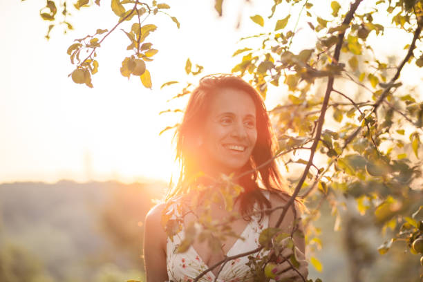 Portrait of smiling, confident mature woman standing along apple tree Beautiful mature woman in white sleeveless dress standing along apple tree during Sunset golden hour stock pictures, royalty-free photos & images