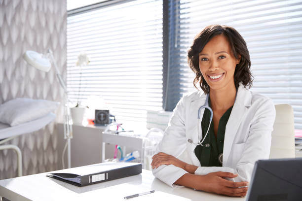 porträt der smiling ärztin, die white coat mit stethoscope-sitting behind desk im büro trägt - allgemeinarztpraxis stock-fotos und bilder