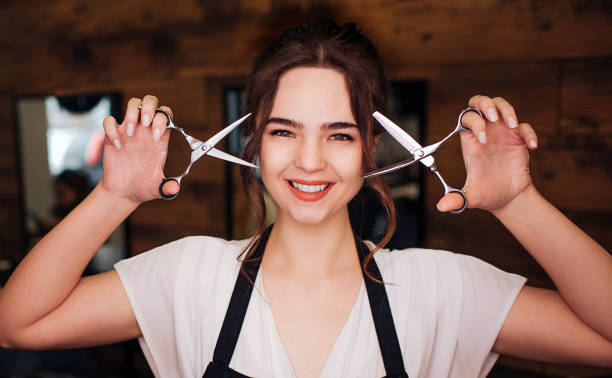 Portrait of smiling hairdresser in beauty salon. Beautiful young woman in black apron holding professional scissors. Beauty and people concept stock photo