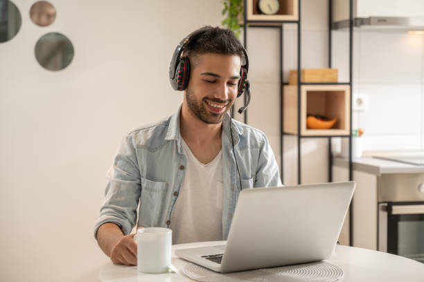 Portrait of smiling man wearing headphones with microphone using laptop at home. stock photo