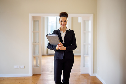 Smiling beautiful saleswoman holding file at empty new apartment. Portrait of female real estate agent. She is wearing blazer.
