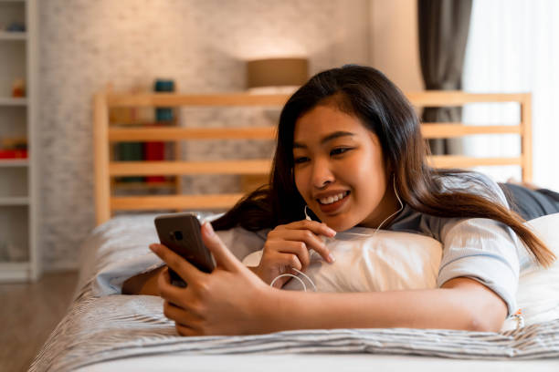 Portrait of smiling young Asian girl lying down on bed while making a video call in bedroom at home. Video conferencing technology and wireless communication concept Portrait of smiling young Asian girl lying down on bed while making a video call in bedroom at home. Video conferencing technology and wireless communication concept. indonesian girl stock pictures, royalty-free photos & images