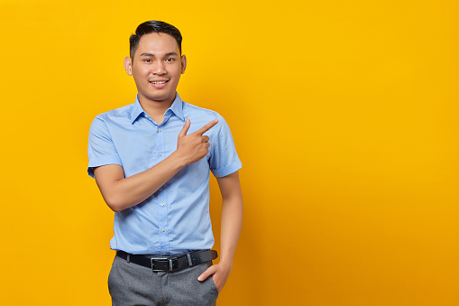 portrait of smiling young asian man in glasses pointing finger away at copy space isolated on yellow background. businessman and entrepreneur concept