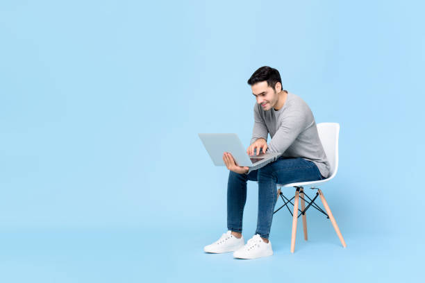 Portrait of smiling young handsome Caucasian man sitting while working on his laptop in isolated studio blue background stock photo