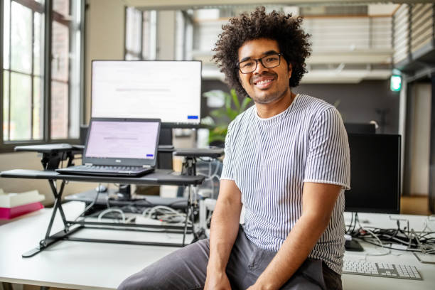 Portrait of smiling young professional sitting at desk Portrait of smiling young professional sitting at desk. Businessman with curly hair sitting on his work desk and looking at camera. 25 29 years photos stock pictures, royalty-free photos & images