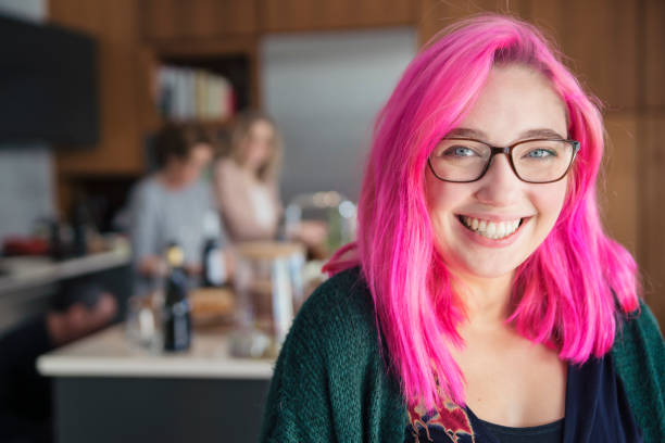 Portrait of smiling young woman with pink hair at big family dinner. Portrait of smiling young woman with pink hair and glasses in home kitchen, while family members are preparing food in the background. Horizontal head and shoulders shot in natural light. Copy space space available. This was shot in Quebec, Canada. pink hair stock pictures, royalty-free photos & images