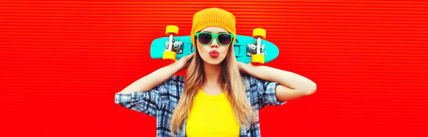 Portrait of stylish blonde young woman model with skateboard wearing colorful yellow hat on red background stock photo